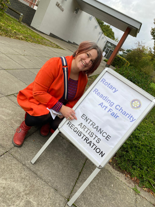 Jeanne-Louise wears a bright orange coat and is crouched next to an A-frame sign for the Reading Charity Art Fair artist registration. The photo is taken outside on the path leading up to the exhibition venue.