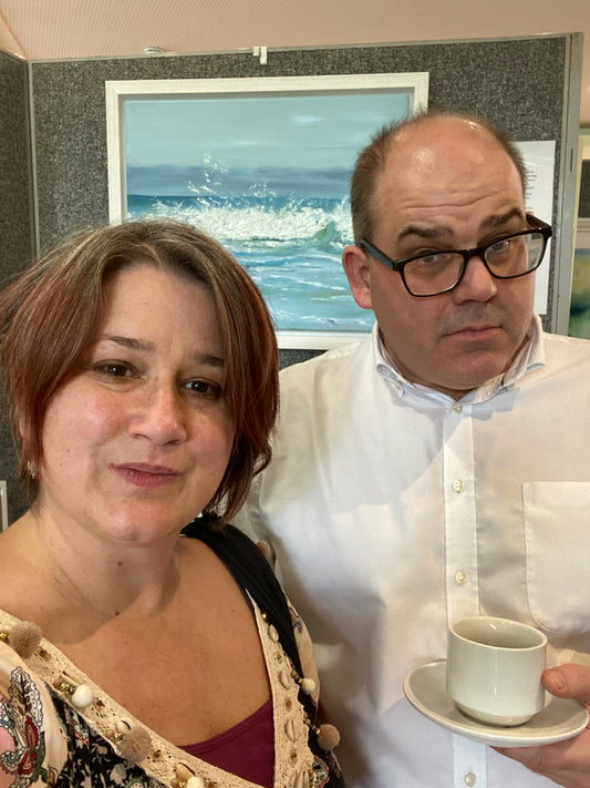 A woman and a man standing in front of an art display with a seascape painting visible. He is holding a coffee cup.