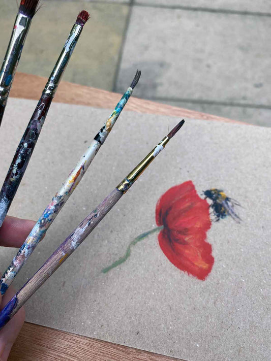 Paint brushes held up above a hand painted greetings card featuring a bee alighting on a red poppy,