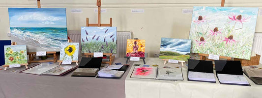 Selection of paintings on table easels displayed at pop-up market with additional artwork in folders on the table in front of the display stands