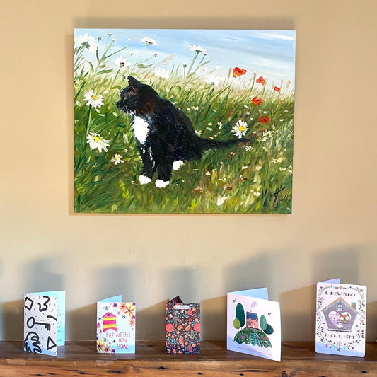 Painting of black and white cat in garden with daisies and poppies hung above a mantelpiece with greetings cards on it