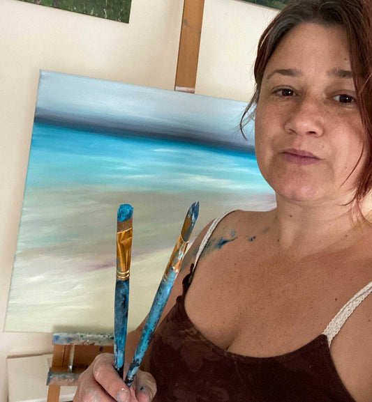 Jeanne-Louise holding paint brushes in front of her easel with a seascape oil painting on it