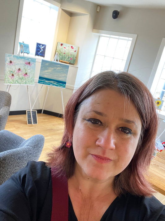 Selfie of Jeanne-Louise in exhibition venue with paintings on easels, shelves and window sills behind her