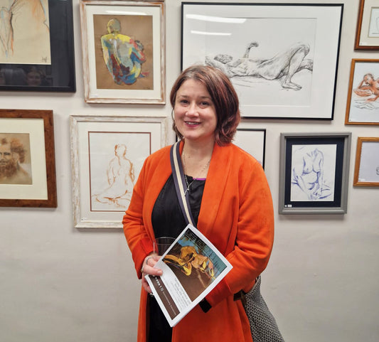 Jeanne-Louise wearing an orange coat stands in front of an exhibition wall with framed life drawings