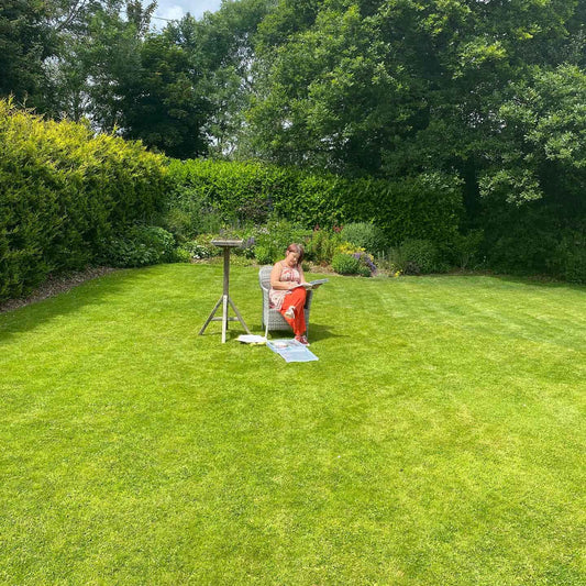 Jeanne-Louise (in orange) on a garden wicker chair placed on a lawn sketching on her lap