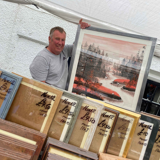 A smiling man holding up a framed artwork in Chinese brush style. He is standing behind a market stall with picture frames for sale.