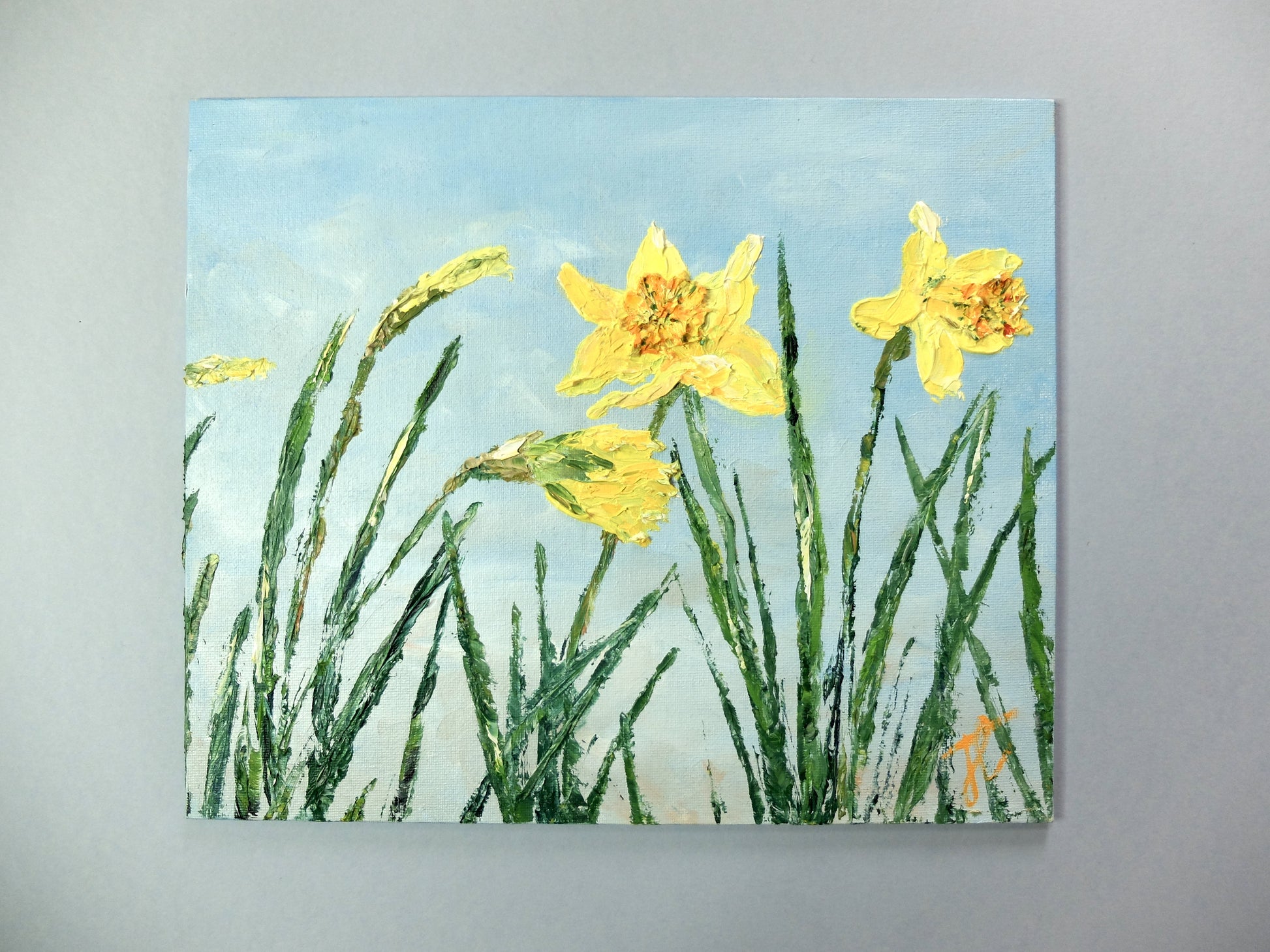 Painting of a row of daffodil plants in bloom, the ones to the left of the painting are in different stages of bud and the two to the right are fully open. The background is a blue sky with soft hints of cloud. The painting has a textured surface where the daffodils are. It is photographed against a grey background